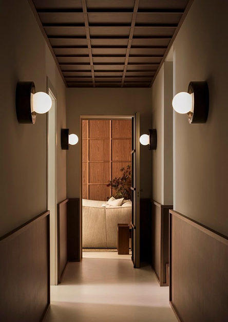 Corridoio minimalista illuminato da moderne applique, che conduce a un accogliente salotto con arredi in legno e divano beige, che incarna uno stile d'interni elegante e rilassante.