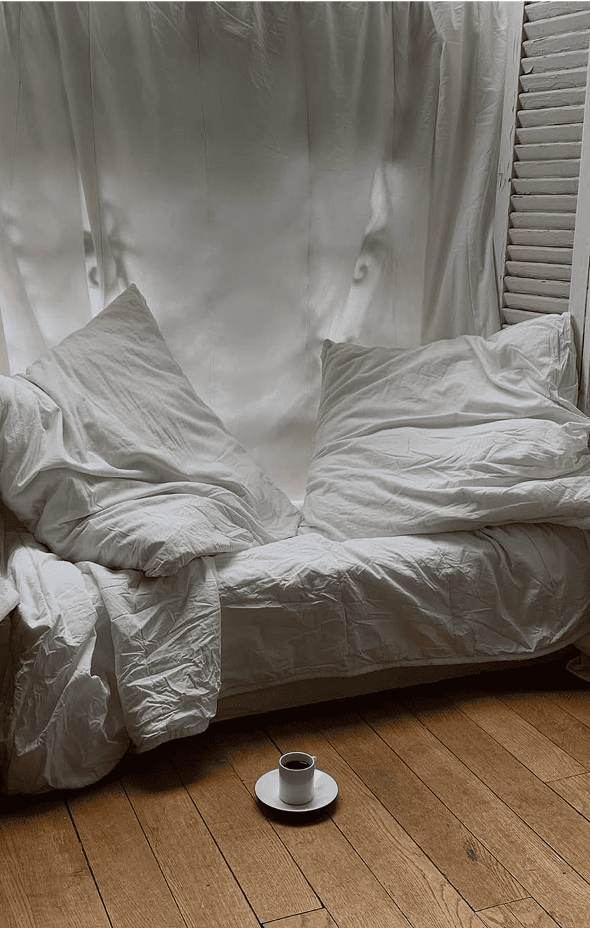 Chambre cosy avec lit défait en draps blancs et tasse de café posée au sol, illustrant une ambiance slow living et un réveil naturel.