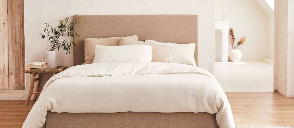 Letto Kipli rivestito in tessuto beige con biancheria chiara in una camera luminosa e naturale