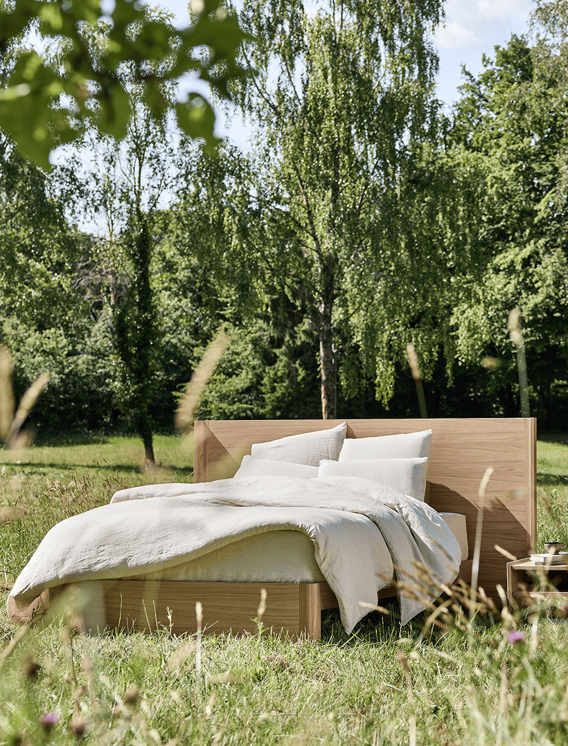 Letto in legno massello Kipli collocato in mezzo alla natura, con biancheria da letto in lino naturale, a illustrare una camera da letto ecologica e un sonno salutare nel cuore di un ambiente verdeggiante.