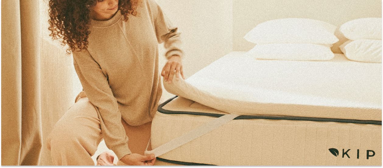 Una donna che sistema un coprimaterasso su un materasso in lattice naturale Kipli in una camera da letto luminosa e minimalista.