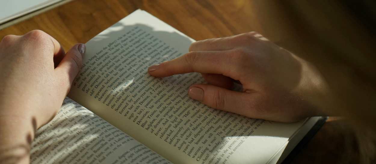 Primo piano sulle mani che sfogliano un libro in un angolo lettura illuminato dalla luce naturale.