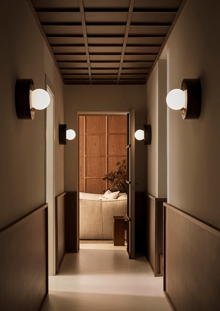 Een minimalistische gang, verlicht door moderne wandlampen, die leidt naar een gezellige woonkamer met houten meubels en een beige bank, wat een elegante en rustgevende interieurstijl weerspiegelt.