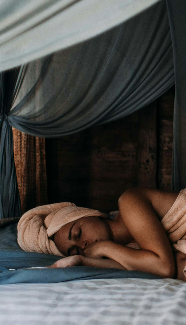 Donna che si riposa dopo un momento di relax, distesa su un letto circondato da tende drappeggiate, che evoca un sonno ristoratore e un’atmosfera di benessere naturale.