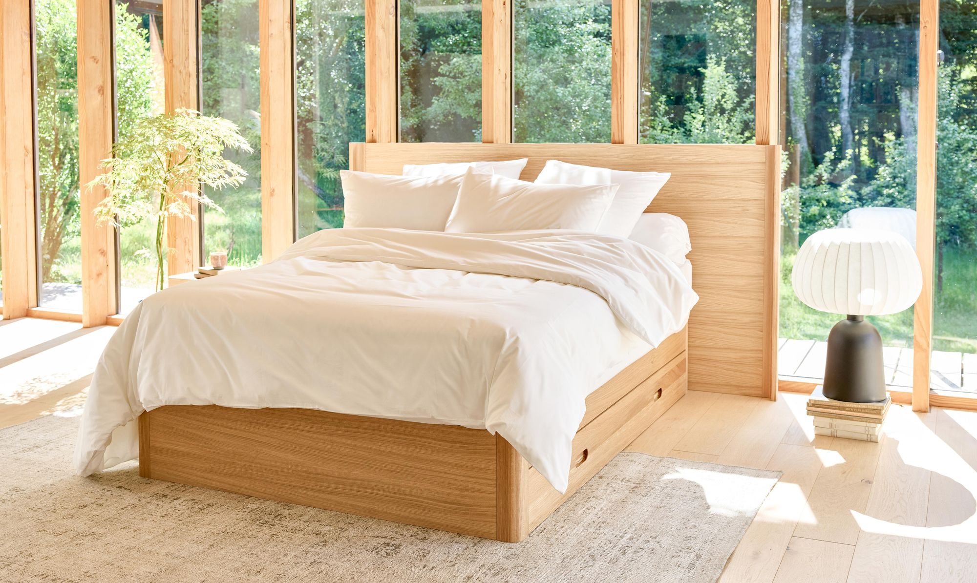 Quarto luminoso com cama em carvalho com gavetas da marca Kipli, lençóis brancos e luz natural que entra pelas grandes janelas panorâmicas.